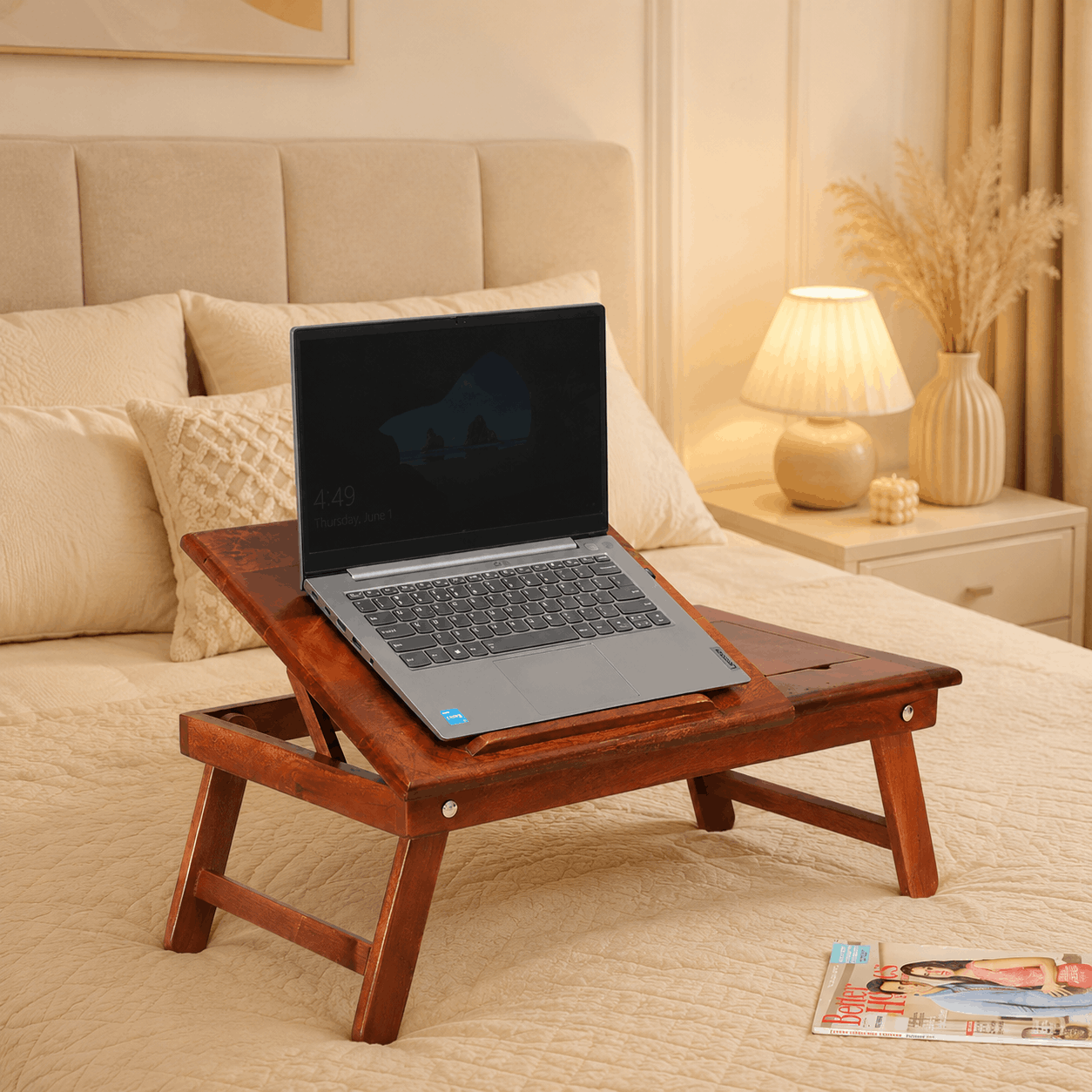 Laptop on a wooden tray on a bed with a lamp and decorative items in the background.