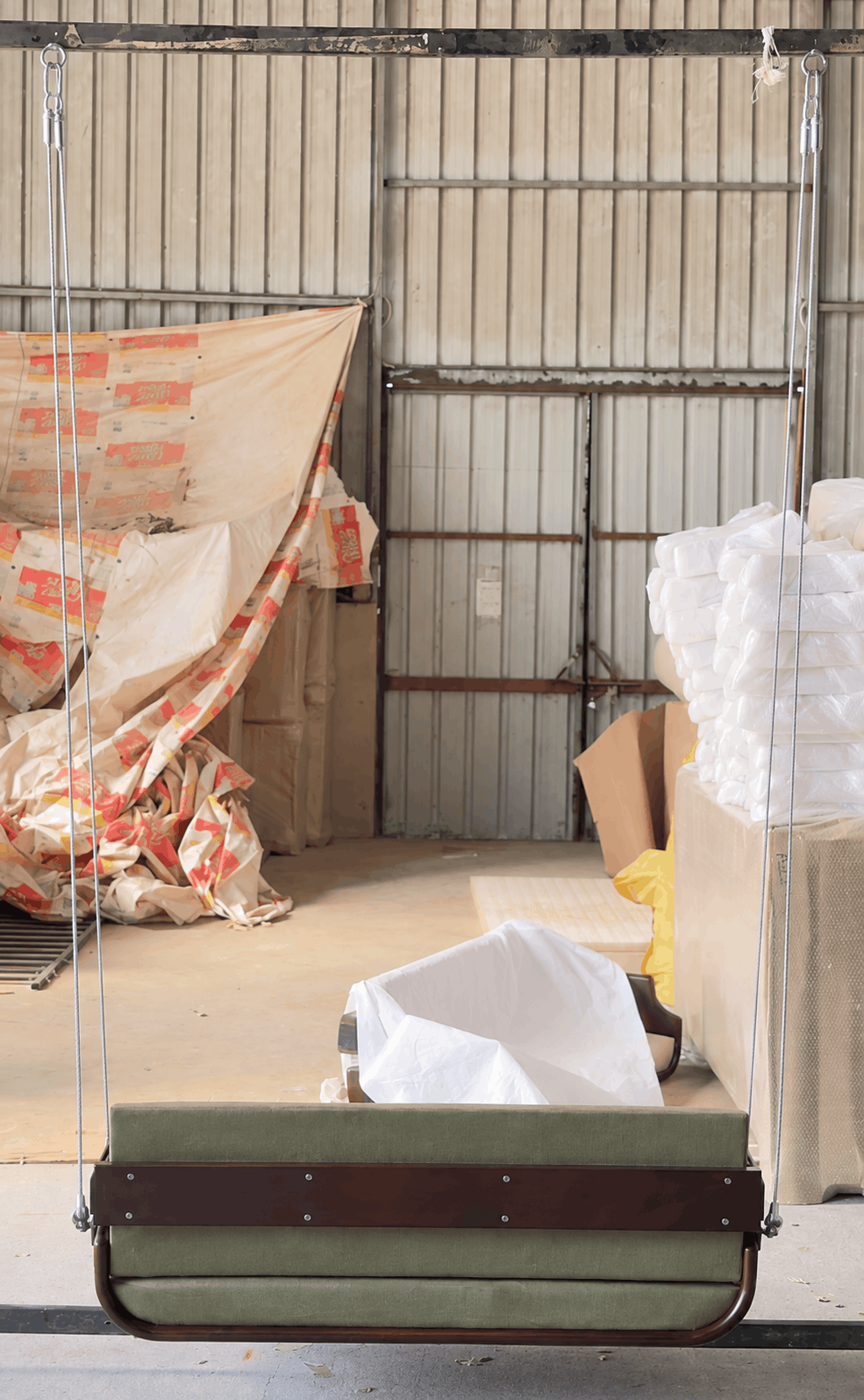 A fabric-covered hanging swing bench with a green cushioned seat and dark brown wooden frame, suspended in a warehouse or storage area with corrugated metal walls and wrapped materials in the background.