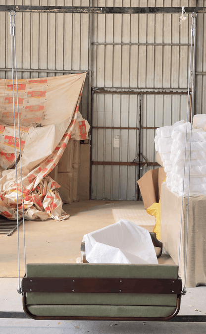 A fabric-covered hanging swing bench with a green cushioned seat and dark brown wooden frame, suspended in a warehouse or storage area with corrugated metal walls and wrapped materials in the background.