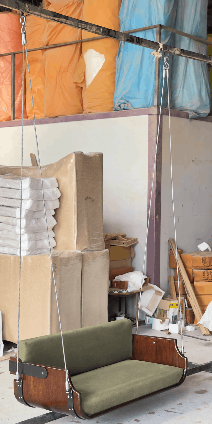 A wooden swing bench with an olive green cushioned seat and backrest, suspended by metal chains in a workshop or warehouse setting with stacked furniture parts and cardboard boxes in the background.