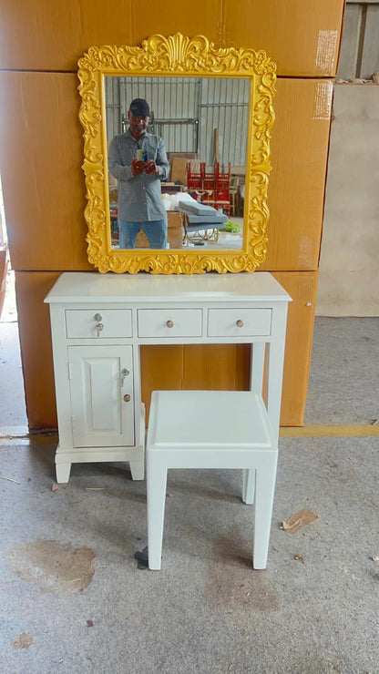White vanity table with a yellow-framed mirror and stool against a wooden wall.